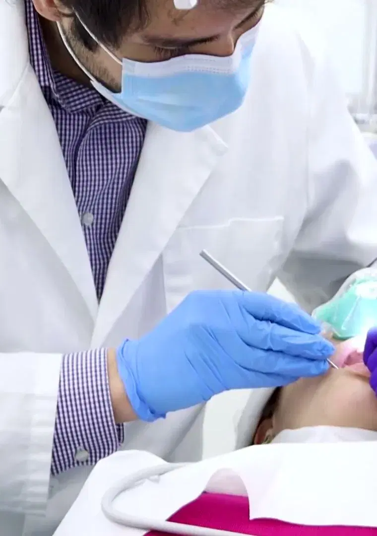 Odontólogo con mascarilla y guantes azules trabajando con un instrumento dentro de la boca de una paciente recostada en el sillón