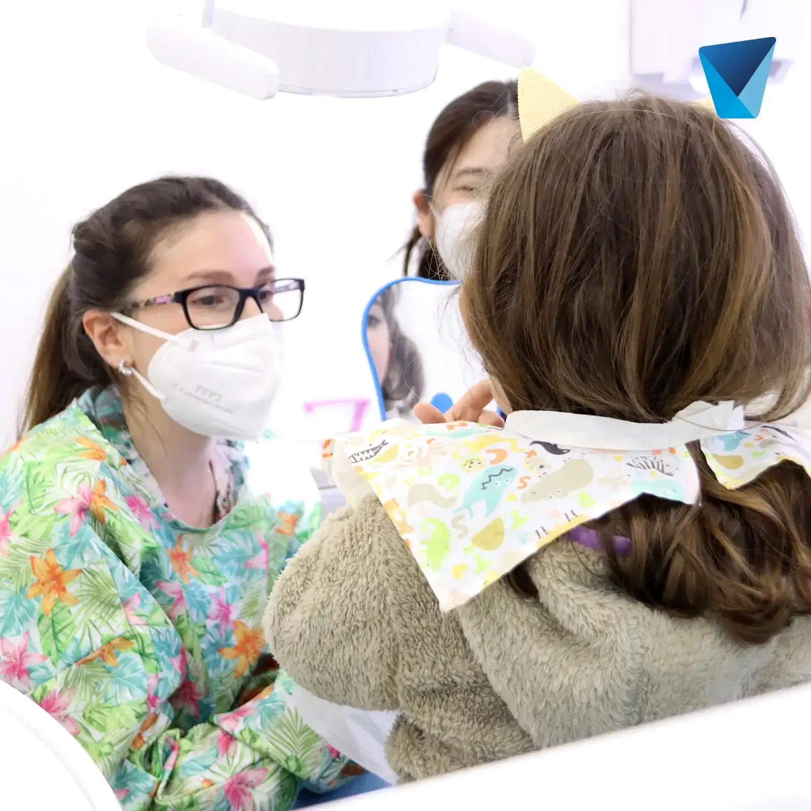 Odontopediatra con mascarilla sentada frente al paciente, que espera con el babero puesto y la asistente detrás, antes de empezar la atención