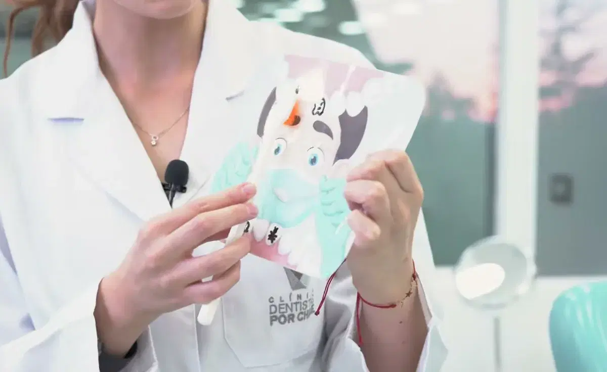 Dentista con delantal blanco sostiene una lámina plastificada con el dibujo de un diente que se cepilla, el material con que la clínica enseña el cepillado a los niños