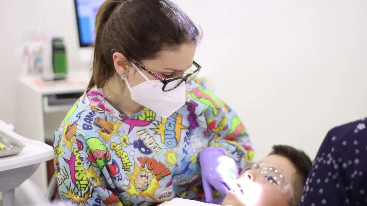 Odontopediatra con mascarilla y guantes aplica la lámpara de fotocurado en la boca de un niño con lentes de protección