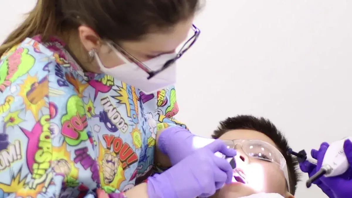 Odontopediatra inclinada sobre un niño con lentes de protección, trabajando en su boca con un instrumento y la luz encendida