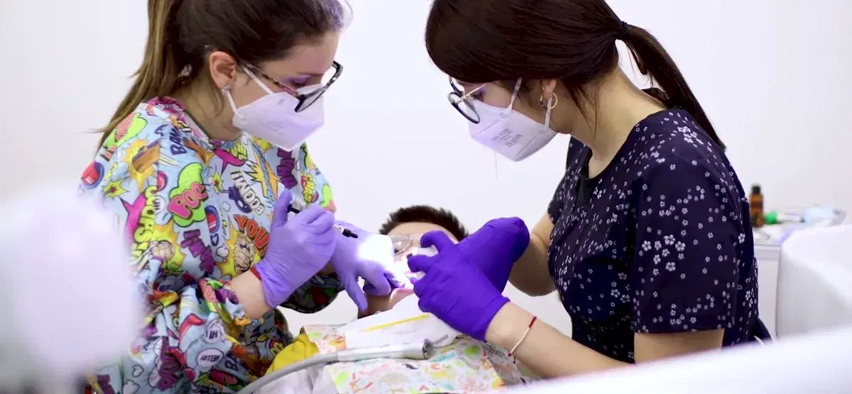 Odontopediatra y asistente, con mascarilla y guantes, trabajando juntas sobre un niño en el sillón con la luz de la lámpara sobre su boca