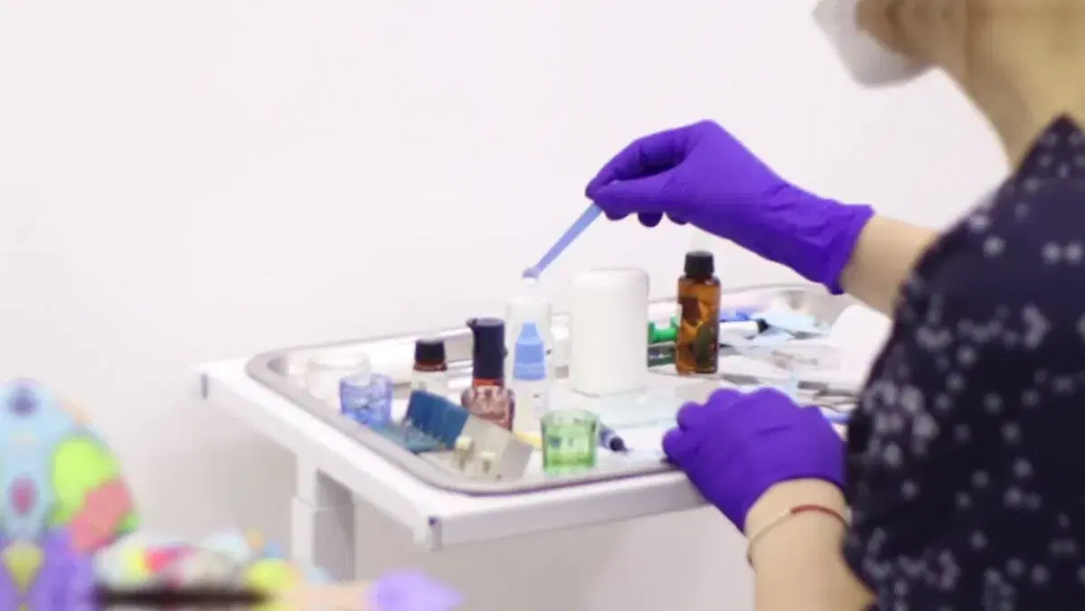 Mano con guante morado sostiene un aplicador sobre la bandeja de materiales dentales, con los frascos y los envases de mezcla ya preparados