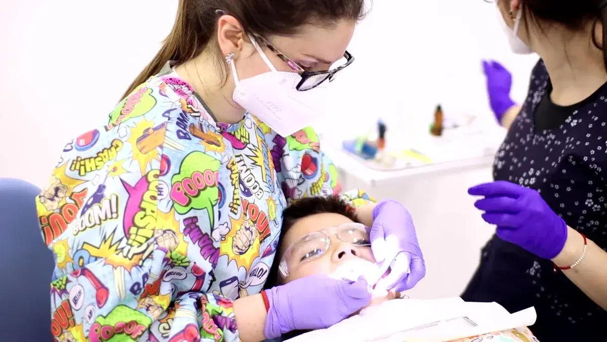 Odontopediatra con mascarilla atiende a un niño recostado en el sillón con lentes de protección, mientras la asistente la acompaña al otro lado