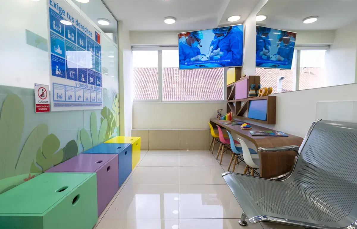 Sala de espera infantil con bancas de colores, mesa de dibujo y pantallas con dibujos animados